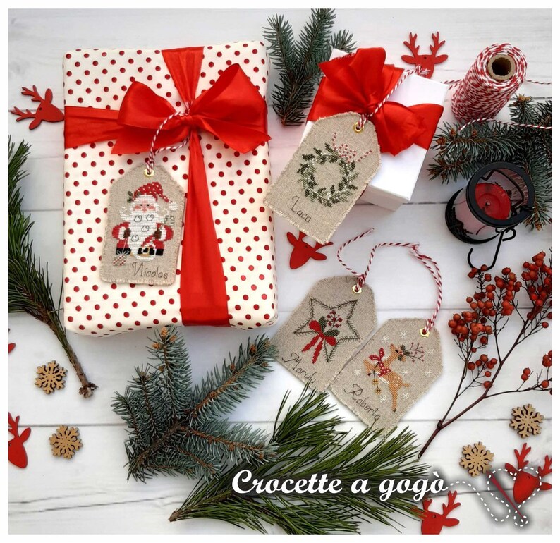 Puede incluir: Cuatro etiquetas de regalo de Navidad con papel de regalo de lunares rojos y blancos, cinta roja y cuerda. Las etiquetas est&aacute;n hechas de arpillera y presentan dise&ntilde;os bordados de Pap&aacute; Noel, una estrella, un reno y una corona. Las etiquetas est&aacute;n etiquetadas con los nombres "Nicolas", "Marie", "Robert" y "Luca".