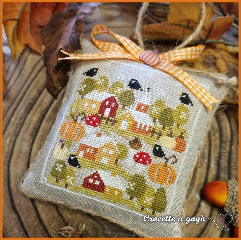Op de afbeelding: Een kruissteek decoratief kussen met een herfstdorp-sc&egrave;ne met huizen, bomen, pompoenen en vogels. Het kussen heeft een jute rand en is versierd met een ruitjeslint en een knoop. De tekst "Croccette a gog&ograve;" is zichtbaar.