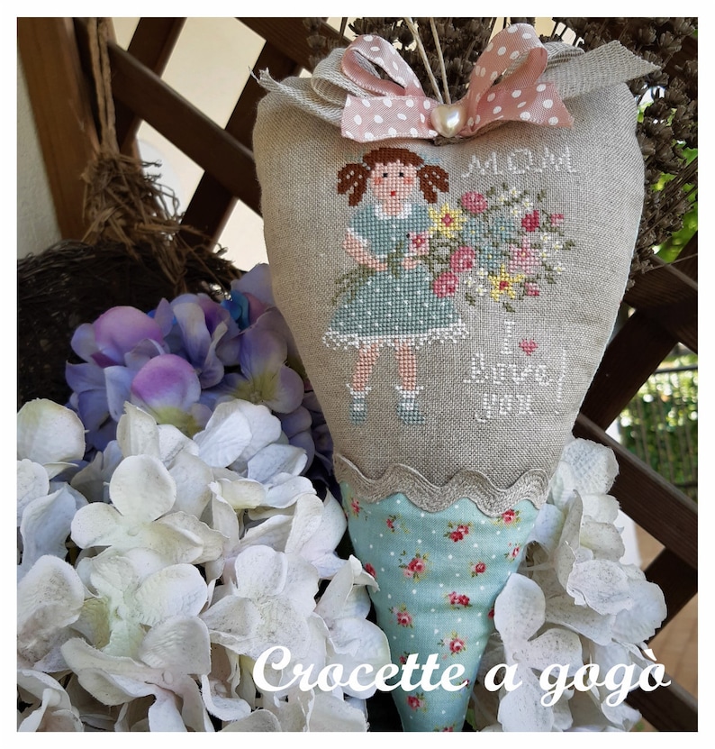 May include: A heart-shaped fabric cone with a stitched image of a girl holding flowers. The cone is decorated with a pink and white polka dot ribbon and a lace trim. The text "Mama I love you" is stitched on the cone. The cone is sitting on a bed of white flowers.