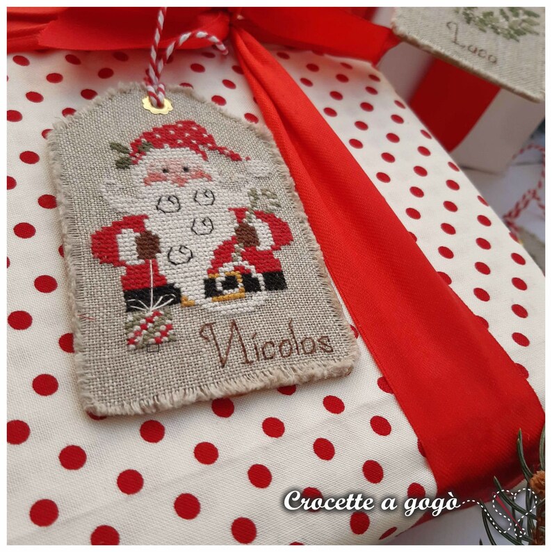 Puede incluir: Una bolsa de regalo blanca con lunares rojos y una cinta roja. Un adorno hecho a mano con punto de cruz con un dise&ntilde;o de Pap&aacute; Noel est&aacute; sujeto a la bolsa. El adorno est&aacute; etiquetado como "Nicolos".