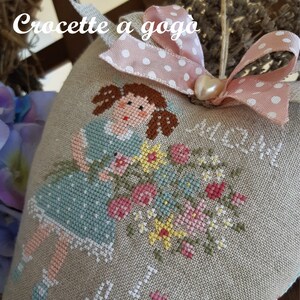 May include: A heart-shaped pillow with a cross-stitch design of a girl holding flowers. The pillow is made of beige linen and has a pink ribbon with white polka dots around it. The text "Crocette a goga" is stitched on the pillow.