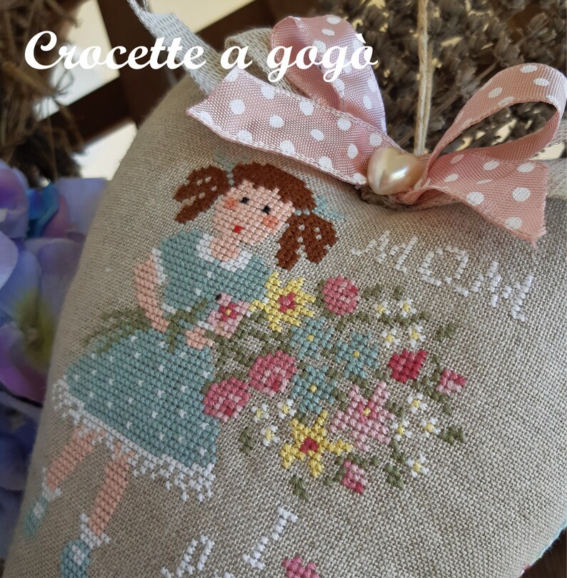 May include: A heart-shaped pillow with a cross-stitch design of a girl holding flowers. The pillow is made of beige linen and has a pink ribbon with white polka dots around it. The text "Crocette a goga" is stitched on the pillow.
