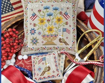 May include: A patriotic needlepoint pillow with a floral design, American flags, and an eagle. The pillow features sunflowers, blue flowers, and stars. A smaller matching piece is also present, along with red, white, and blue ribbons and a pair of gold scissors.