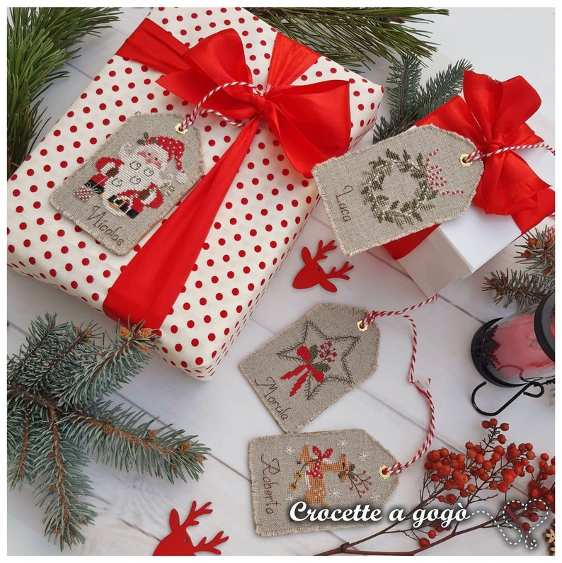 Puede incluir: Cuatro etiquetas de regalo de Navidad hechas a mano con cuerda roja y blanca. Las etiquetas est&aacute;n hechas de tela de lino natural y presentan dise&ntilde;os de punto de cruz con nombres, incluyendo "Nicholas", "Luca", "Marla" y "Roberta". Las etiquetas est&aacute;n atadas con cuerda roja y blanca y est&aacute;n listas para ser adjuntadas a los regalos.