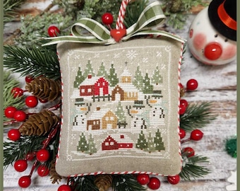 May include: A beige fabric Christmas ornament with a red and white striped ribbon and a green and white plaid bow. The ornament features a cross-stitch design of a snowy village with houses, trees, and snowmen. The text "Crocette a gogo" is printed on the bottom of the ornament.