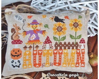 May include: A cross-stitch pillow featuring a colorful autumn scene with a scarecrow, sunflowers, mushrooms, a squirrel, and the word "AUTUMN" in large letters. The pillow is made of beige fabric and has a brown border.