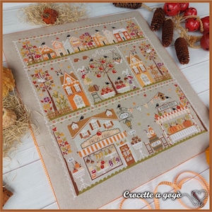 May include: Cross-stitch pattern featuring a charming autumn village scene with houses, trees, pumpkins, and a "Fresh Fruit" shop. The pattern is on a beige linen fabric with orange stitching around the edges. The text "Crocette a gogo" is visible in the bottom right corner.