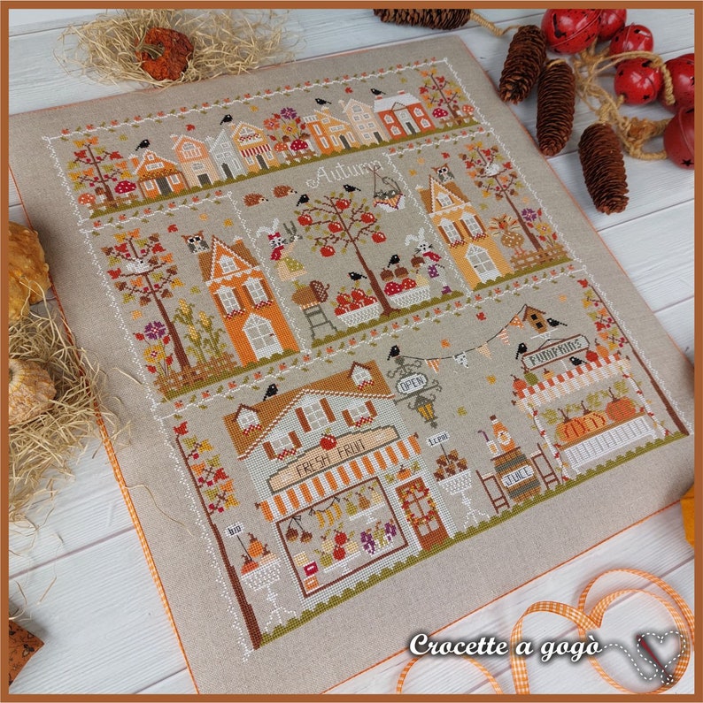May include: Cross-stitch pattern featuring a charming autumn village scene with houses, trees, pumpkins, and a "Fresh Fruit" shop. The pattern is on a beige linen fabric with orange stitching around the edges. The text "Crocette a gogo" is visible in the bottom right corner.