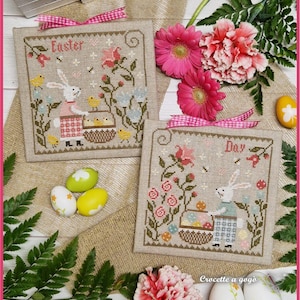 Può includere: Due opere d'arte a punto croce a tema pasquale, con disegni floreali e di conigli. Una recita "Easter" e l'altra "Day". Nastri e fiori rosa circondano le opere, con uova di Pasqua decorative e verde.