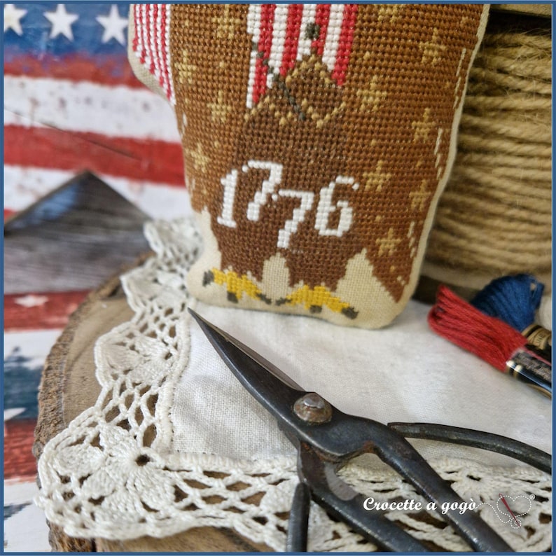 May include: A handcrafted, patriotic needlepoint piece featuring an eagle design and the year "1776" in white. The piece is brown, beige, red, and blue. Vintage scissors and embroidery floss are also visible, suggesting a crafting theme.
