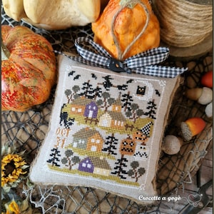 Può includere: Un ornamento quadrato in tessuto beige con una scena di Halloween a punto croce raffigurante case, alberi, un gatto nero, un gufo e il testo OCT 31. È completato da un fiocco in nastro a quadretti bianco e nero e un cappio in iuta.