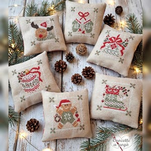 May include: Six square beige fabric ornaments featuring cross-stitch designs of a reindeer, mittens, a bell, a mug, a gingerbread person, and a stocking. Each ornament is bordered by a decorative stitch and includes the text Crocette a gogò.