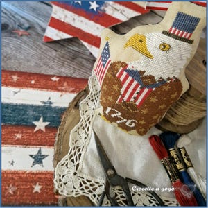 May include: A patriotic needlepoint eagle with a top hat and American flag, featuring the year "1776". The eagle is set on a white lace doily, with red, white, and blue embroidery floss and vintage scissors. A wooden star and flag are in the background.