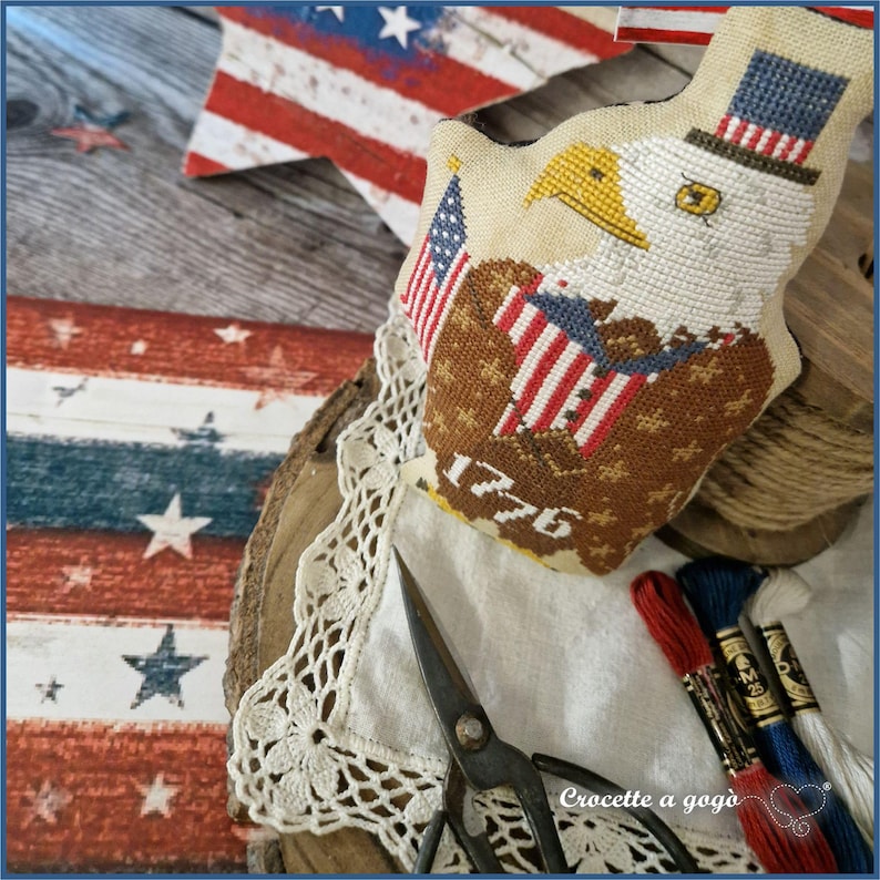 May include: A patriotic needlepoint eagle with a top hat and American flag, featuring the year "1776". The eagle is set on a white lace doily, with red, white, and blue embroidery floss and vintage scissors. A wooden star and flag are in the background.