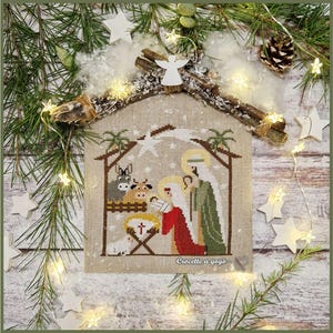 May include: A cross-stitch depiction of the Nativity scene, featuring Mary, Joseph, and baby Jesus in a stable. The artwork is framed by a rustic wooden structure and adorned with stars, an angel, and festive lights. The text "Crocette a gogo" is visible.