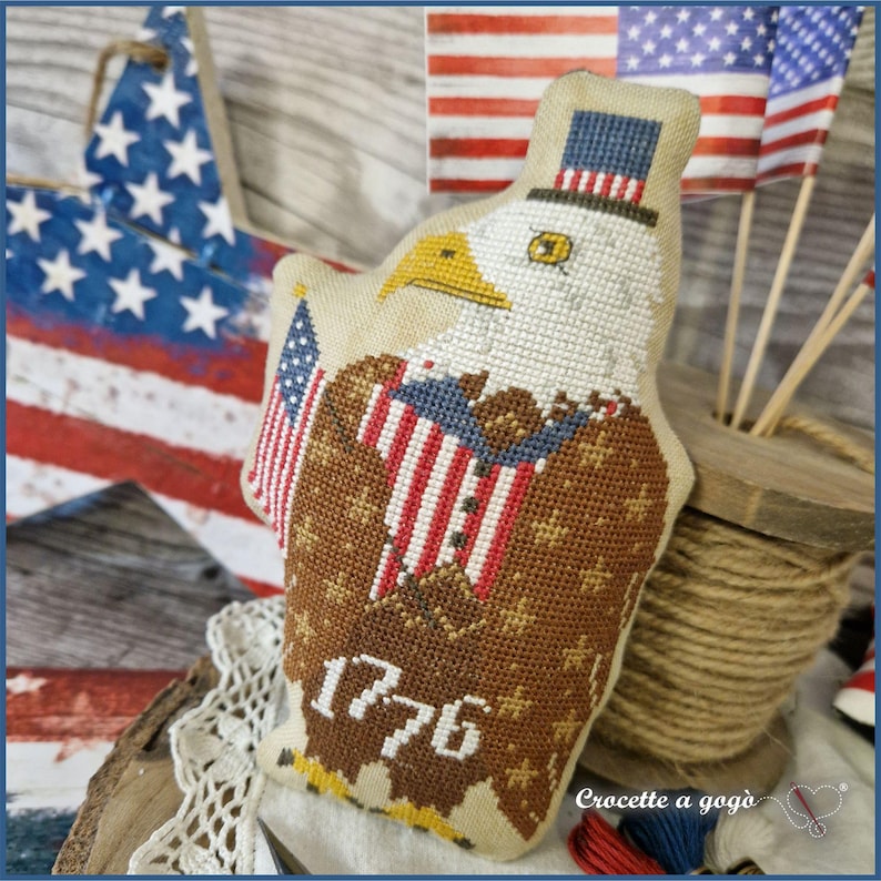 May include: A patriotic, cross-stitched eagle ornament with a top hat and American flag details. The eagle features brown, white, red, and blue colors, with the year "1776" stitched on the bottom. The background includes American flags and a wooden star.