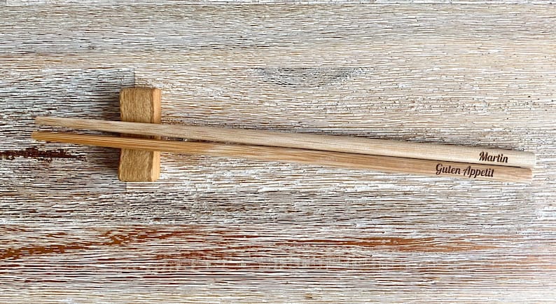 K&ouml;nnte beinhalten: Ein Paar Holzst&auml;bchen mit den Worten "Martin" und "Guten Appetit" eingraviert. Die St&auml;bchen liegen auf einem Holzst&auml;bchenhalter.