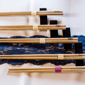 K&ouml;nnte beinhalten: Vier Paar Holzst&auml;bchen mit eingravierten Namen, die auf einem blauen Spitzenstoff und schwarzen St&auml;ndern liegen. Die Namen sind Esther, Steffen, Jens und Ines.