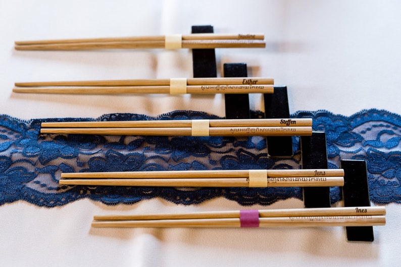 K&ouml;nnte beinhalten: Vier Paar Holzst&auml;bchen mit eingravierten Namen, die auf einem blauen Spitzenstoff und schwarzen St&auml;ndern liegen. Die Namen sind Esther, Steffen, Jens und Ines.