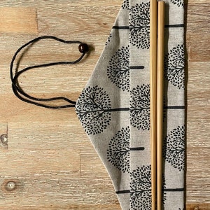 K&ouml;nnte beinhalten: Ein Paar Holzst&auml;bchen in einer beigefarbenen Stofftasche mit einem schwarzen Kordelzug und einem schwarzen und wei&szlig;en Baummuster.