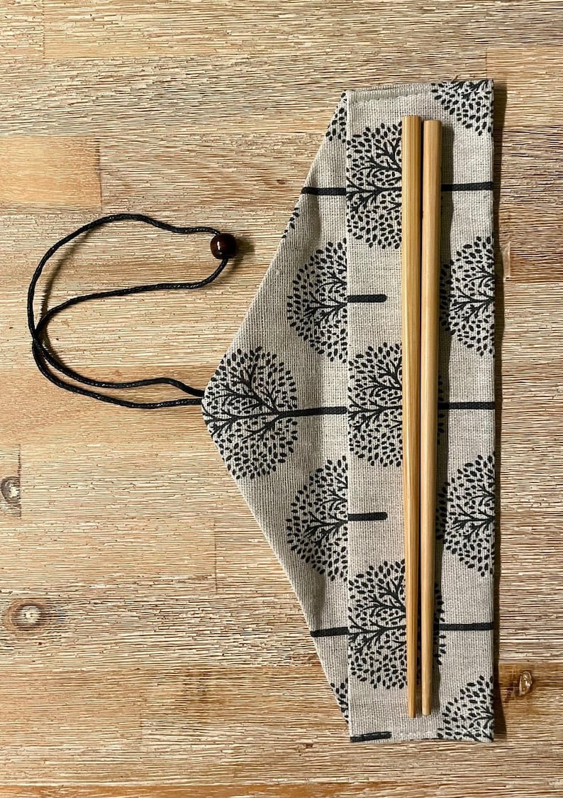 K&ouml;nnte beinhalten: Ein Paar Holzst&auml;bchen in einer beigefarbenen Stofftasche mit einem schwarzen Kordelzug und einem schwarzen und wei&szlig;en Baummuster.