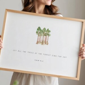 May include: A framed art print featuring a watercolor illustration of several trees. The artwork is set against a white background, with the text "LET ALL THE TREES OF THE FOREST SING FOR JOY" and "PSALM 96:12" below the trees. The frame is made of light-colored wood.