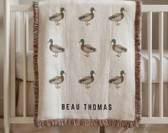 May include: A cream-colored baby blanket with a repeating pattern of watercolor ducks. The blanket has a fringed edge with brown, red, and cream threads. The name "BEAU THOMAS" is printed in black block letters on the blanket.