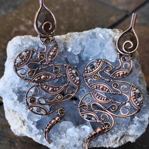 May include: Two snake-shaped wire-wrapped pendants crafted from copper wire, featuring intricate swirling patterns and small embedded dark brown gemstones. Each snake head is accented with a larger, teardrop-shaped dark brown stone.