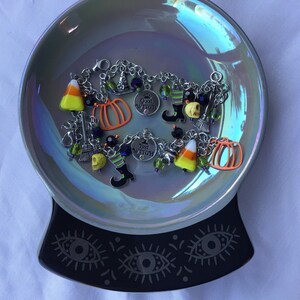 May include: A silver charm bracelet with Halloween themed charms, including a pumpkin, a witch's hat, a candy corn, a skull, and a broom. The charms are on a silver chain with a lobster clasp.