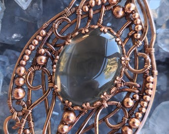 May include: A copper wire wrapped pendant with a clear gray gemstone in the center. The pendant is decorated with copper beads and wire loops.