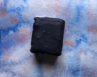 May include: A solid black rectangular watercolor paint pan centered on a square swatch of paper featuring a blended wash of blue and soft brown pigments. The paint pan has a smooth, matte surface and sits directly atop the textured paper background.