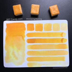 May include: A watercolor paint pan in the color Candy Corn, with a swatch of the color on a white card. The swatch shows the color in different shades and intensities. The card also has the text "007 Candy Corn" and "Shimmering/Staining".
