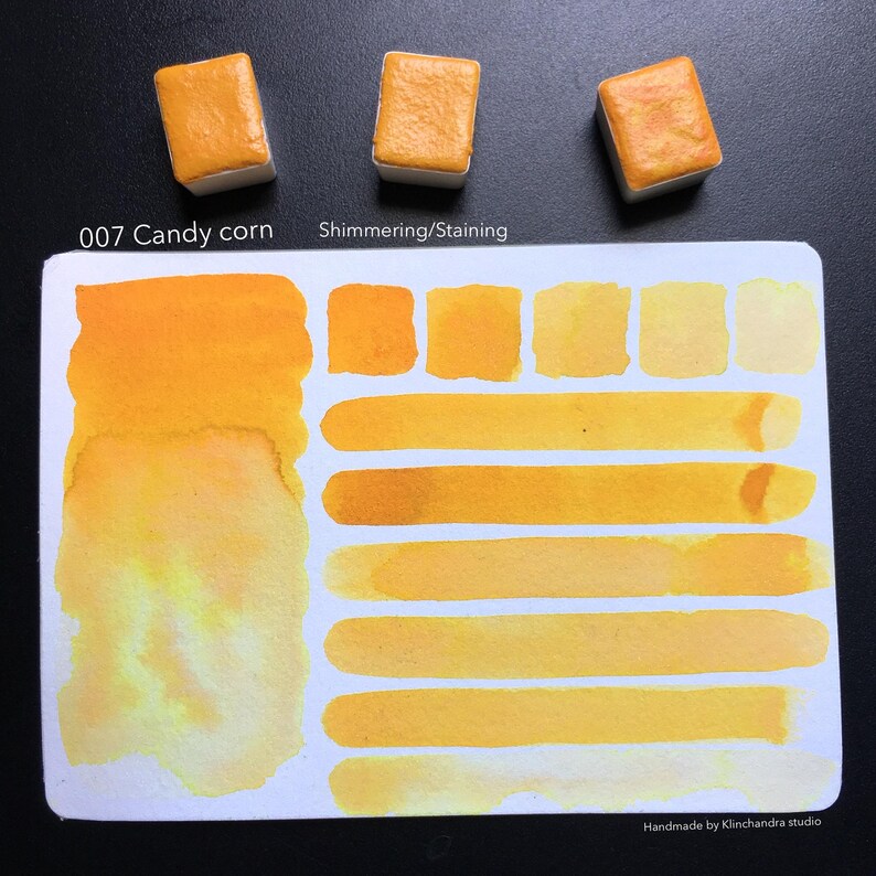 May include: A watercolor paint pan in the color Candy Corn, with a swatch of the color on a white card. The swatch shows the color in different shades and intensities. The card also has the text "007 Candy Corn" and "Shimmering/Staining".