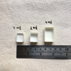 May include: Three white plastic paint pans of different sizes, labeled 1 ml, 2 ml, and 4 ml. A ruler is placed below the pans for size reference.