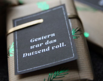 Könnte beinhalten: Geschenke in Kraftpapier verpackt, verziert mit grünen Blättern und einem schwarzen Etikett. Auf dem Etikett steht "Gestern war das Dutzend voll." Eine rote Christbaumkugel ist im Hintergrund zu sehen.