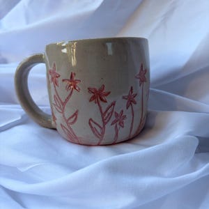 May include: A beige ceramic mug with a handle, adorned with a hand-drawn floral design in red. The flowers and stems are delicately sketched around the mug's exterior, creating a charming, handcrafted aesthetic. The mug is set against a white background.