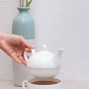 May include: A white porcelain teapot is held above a white teacup filled with tea, on a saucer. A light blue and white vase with greenery is in the background. The scene is set on a light-coloured surface.