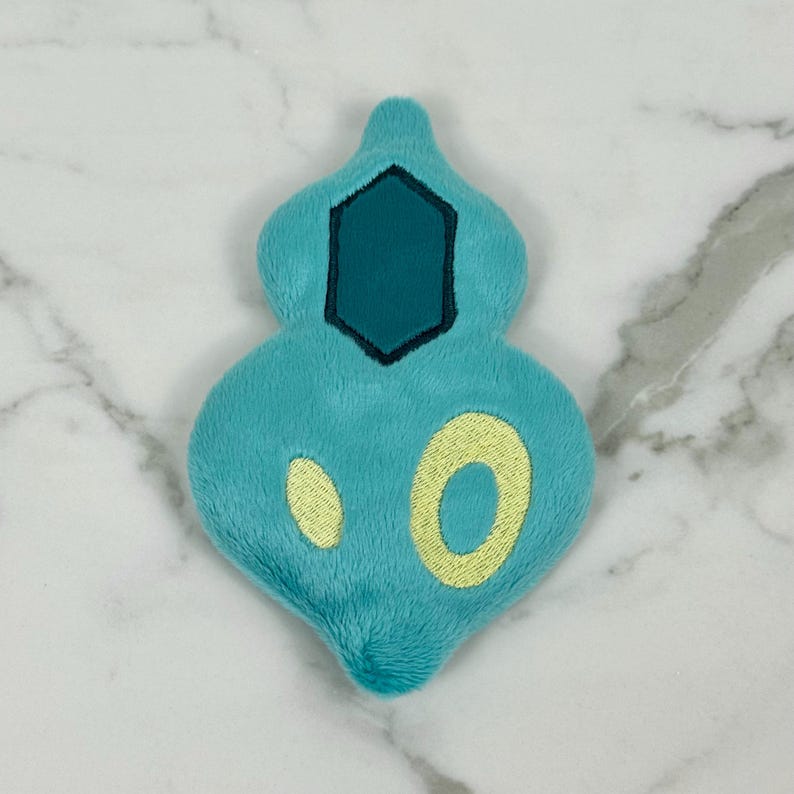 May include: A plush, teal-colored toy in a unique shape, featuring a dark teal hexagon and two yellow ovals. The toy is made of soft material and is likely a character from a popular franchise.