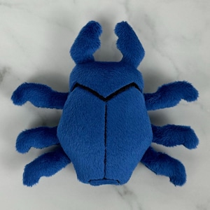 May include: A blue plush toy shaped like a beetle with six legs. The beetle has a black embroidered design on its back.