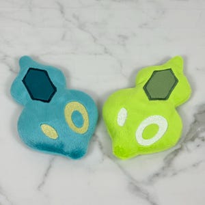 May include: Two plush toys in the shape of a stylized teardrop. One is light blue with a dark blue hexagon and yellow circle. The other is neon green with a dark green hexagon and white circle. Both have a soft, fuzzy texture.