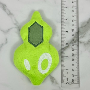 May include: A lime green plush toy with a geometric design and white accents. The toy is approximately 3 inches long. A ruler is placed next to the toy for scale.