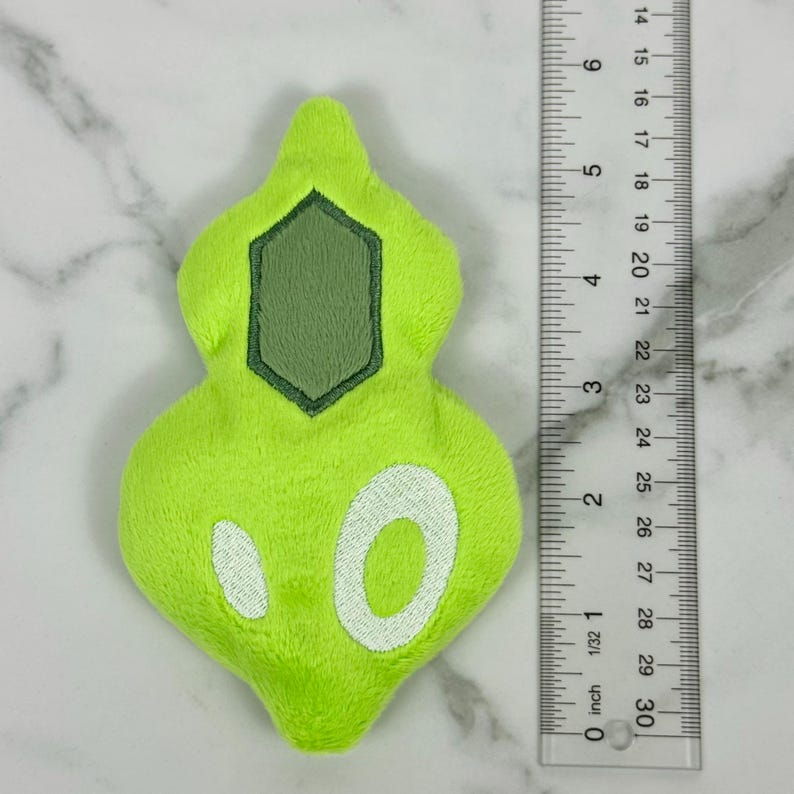 May include: A lime green plush toy with a geometric design and white accents. The toy is approximately 3 inches long. A ruler is placed next to the toy for scale.