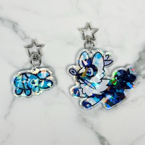 May include: Two iridescent keychains with star-shaped metal clasps. One keychain features a blue and white cartoon animal with a black nose and eyes. The other keychain features a blue and white cartoon animal with a black nose and eyes.