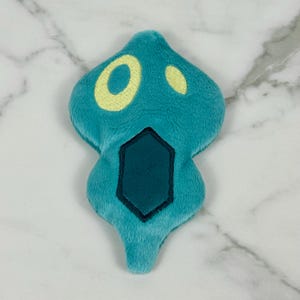 May include: A teal plush toy shaped like a stylized ghost or spirit. It features a yellow eye and a smaller yellow dot, with a dark teal geometric shape at the bottom. The toy is made of soft, fuzzy material.