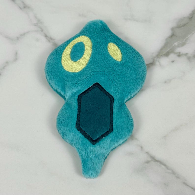 May include: A teal plush toy shaped like a stylized ghost or spirit. It features a yellow eye and a smaller yellow dot, with a dark teal geometric shape at the bottom. The toy is made of soft, fuzzy material.