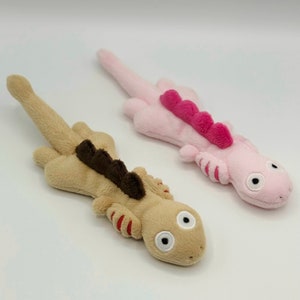 May include: Two plush axolotl toys. One is beige with a brown dorsal fin and red accents on its limbs. The other is pink with a fuchsia dorsal fin and pink accents. Both have large, round eyes and are lying down.