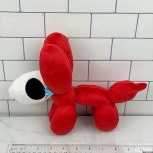 May include: A plush toy shaped like a red balloon dog with a white head. The dog has black eyes and a blue tear. A ruler is placed near the toy. The toy is approximately 10 inches long.