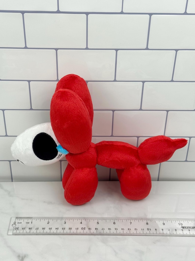 May include: A plush toy shaped like a red balloon dog with a white head. The dog has black eyes and a blue tear. A ruler is placed near the toy. The toy is approximately 10 inches long.