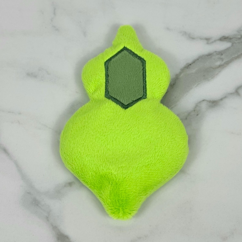 May include: A lime green plush toy in a unique, abstract shape. The toy has a darker green, geometric patch in the center. The plush material is soft and fuzzy, and the toy is set against a white marble background.