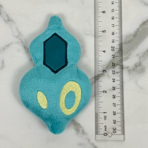 May include: A plush, turquoise-colored figure with a dark teal hexagon and yellow circular accents. The figure is approximately 3 inches long, as measured by the ruler in the image. The plush has a soft, textured appearance.